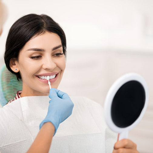 Dentist holding a veneer in front of a patient's smile while she looks in a mirror